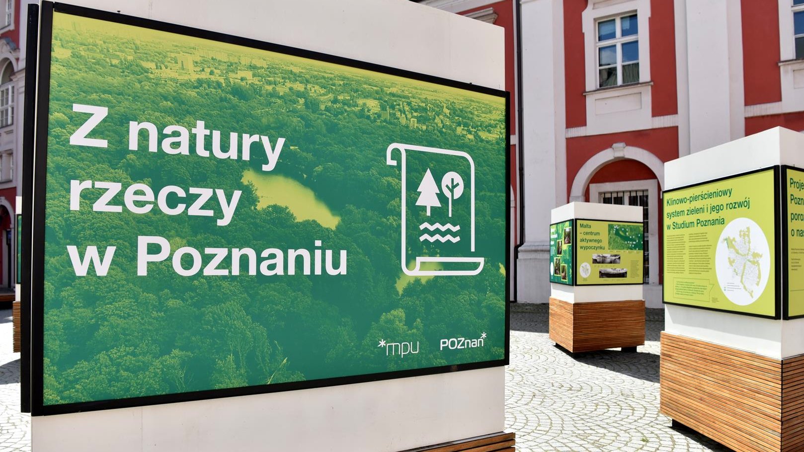 Na zdjęciu plansza wystawy z napisem: z natury rzeczy w Poznaniu. W tle budynek urzędu