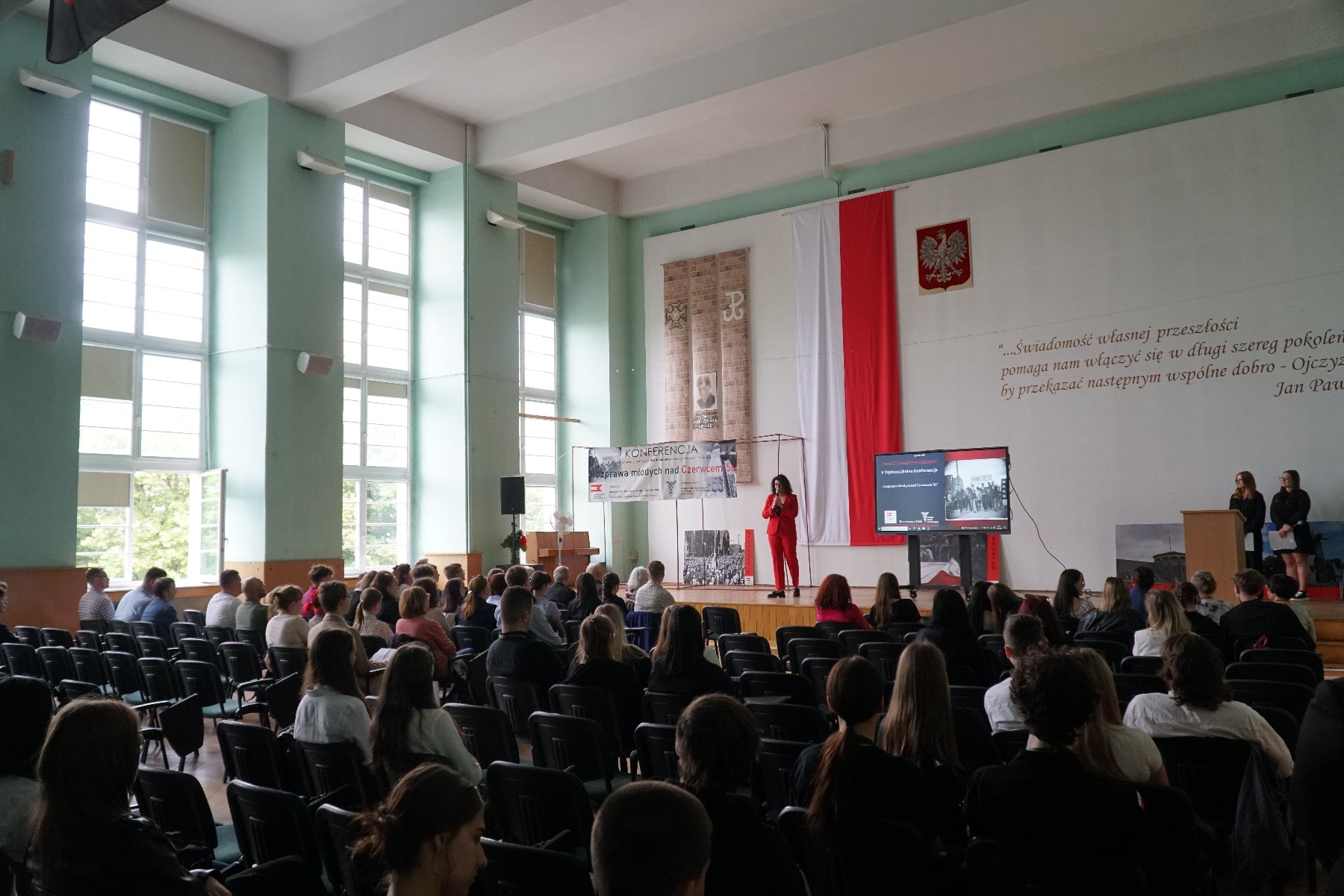 Sala konferencyjna z dużymi oknami. Publiczność siedzi na krzesłach. Na scenie stoi kobieta w czerwonym garniturze, przemawia do uczestników. Za nią na ścianie duża flaga Polski, godło oraz cytat Jana Pawła II. Obok ekran z prezentacją. Po lewej baner z napisem "Konferencja - Wyprawa młodych nad Czerwiec'